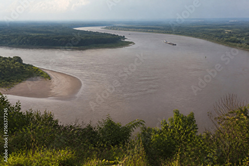 View from high bluff of wide brown river bending through low green forest, sandbar, tugboat and barge downstream, Mississippi River landscape