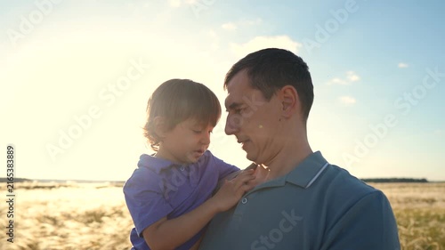 A man holds a boy closely, their faces sharing a tender moment. The boy gazes at the man, who smiles gently, in a sunlit field.