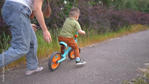 A woman helps a boy ride a blue bike as they move along a path. The boy, focused on biking, is assisted by the woman in a natural setting.