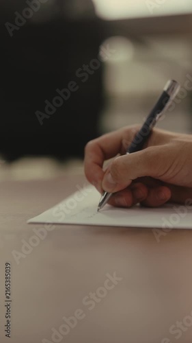 Male hand holding pen and writing on white sheet of paper placed on wooden desk, closeup vertical cropped shot, slow motion. Concept of act of signing contract, taking notes, composing letter.