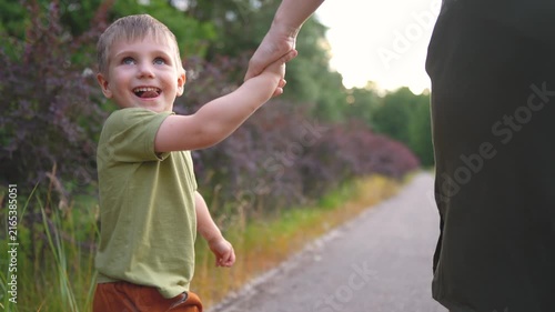 A woman holds a boy's hand as they walk along a path. The boy looks back, enjoying the nature around them and the woman s companionship.