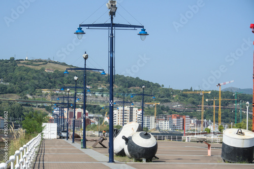 Cuadro en lienzo Blue lamp posts along a quayside promenade with old mooring buoys and green hill