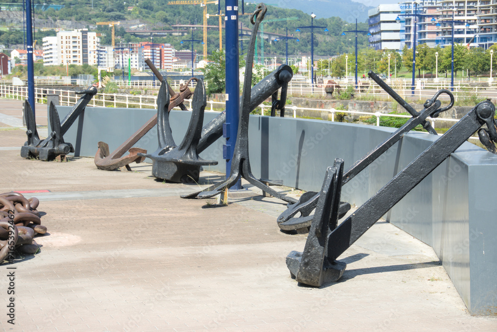 Fototapeta premium Row of historic ship anchors along a quay wall with tower cranes behind