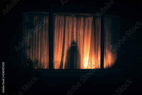 A ghostly figure with glowing eyes stands behind the curtains of an open window in a bedroom at night