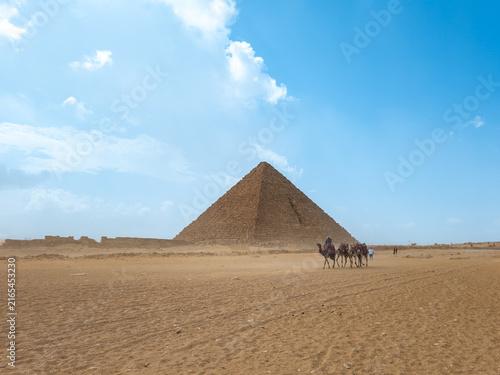 Camels carrying people traveling across the desert plains with the ancient giza pyramid towering in the background