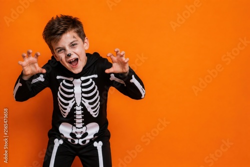 Scared boy in skeleton costume making monster claws on Halloween orange background with copy space