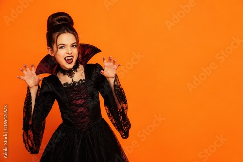 Young girl in vampire costume with fangs and dark dress on orange Halloween orange background with copy space