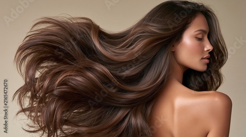 A woman with long, healthy hair captured from behind, gently tossing her hair in slow motion against a soft neutral studio backdrop