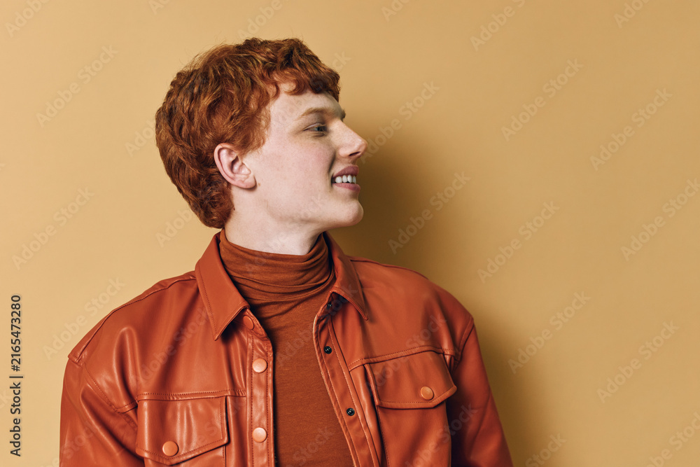 Obraz premium Young man with red hair wearing a brown leather jacket and turtleneck looking sideways smiling against a beige background in studio portrait.