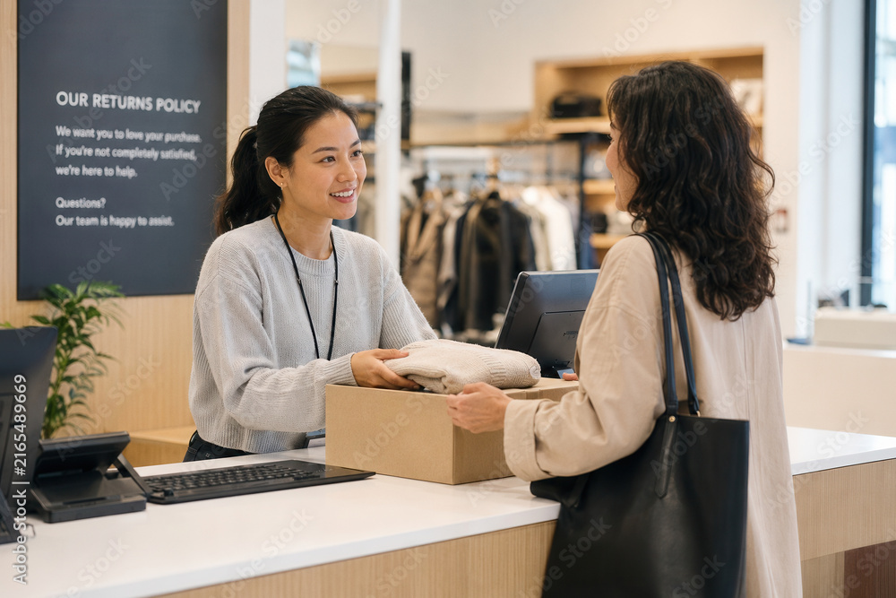 Naklejka premium Retail clerk hands a boxed item back to a customer at a store counter while smiling and speaking by the register near a returns policy sign.