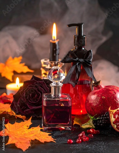 A sophisticated perfume bottle and a red dispenser, adorned with a black ribbon, sit amidst a dramatic display of a single black rose, burning candles, colorful autumn leaves, and juicy pomegranates.