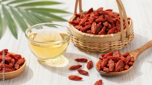 A serene display of dried goji berries in a bamboo basket and wooden spoon, accompanied by a glass cup of golden herbal tea on a white wooden surface with green leaves.