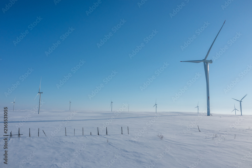 Fototapeta premium Close drone perspective of modern wind turbines standing tall across a vast, pristine snow-covered field under a clear blue sky.