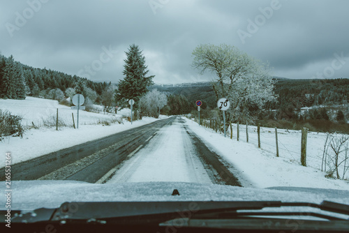 Snow covered rural road viewed through a car windshield in Auvergne France winter forest. A journey through frost laden countryside under heavy grey sky captures driving in harsh conditions.