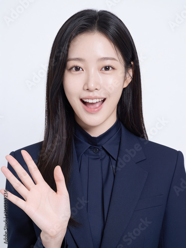 Beautiful young East Asian woman in a dark navy suit waving her hand with a bright cheerful smile in a studio