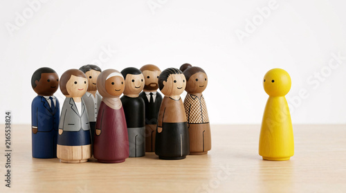 A group of diverse wooden figures face a solitary yellow figure, representing a scenario of inclusion and distinction or perhaps leadership and conformity.