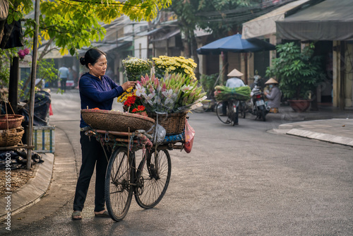 Vietnamese Flower Seller Arranging Fresh Blossoms in Woven Baskets on a Bicycle at an Early Morning Local Neighborhood Market