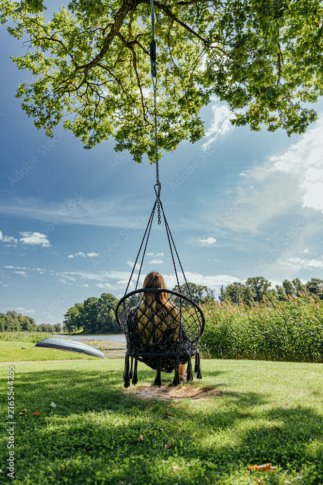 Fototapeta premium Woman seen from behind sits in a hanging chair beneath a leafy tree, overlooking a calm lake with reeds, green lawn and blue summer sky