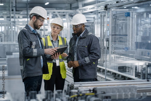 Cross Functional Manufacturing Team Collaborating with a Tablet beside an Automated Production Line in a Clean Modern Industrial Factory