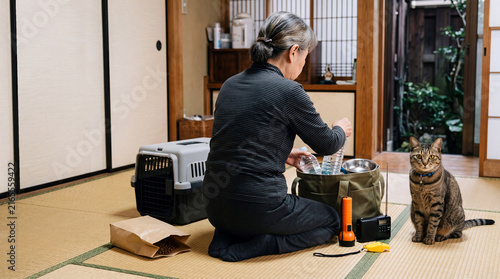猫と暮らす高齢女性のペット防災準備、懐中電灯やラジオを備えた避難用バッグ / Senior woman preparing pet disaster supplies with her cat, an evacuation bag equipped with a flashlight and radio