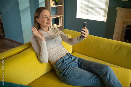 Young woman relaxing on a yellow sofa at home, communicating with friends or family during a video call on her smartphone, sharing moments and staying connected