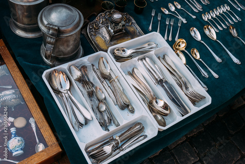 Photography Assorted antique silver spoons, forks, and knives in a white tray container with pewter tankards and metal tableware on a flea market table