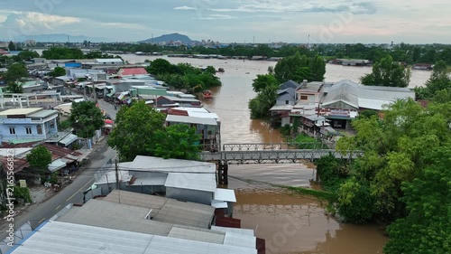 Drone view of a riverside community in An Giang, Vietnam, with brown floodwater surrounding homes, roads, trees, and buildings during the annual wet season.