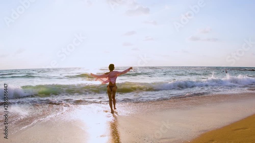 The young slender woman in a stylish swimsuit and shirt walks on the sandy beach, looking at the calm ocean horizon with a sense of freedom and tranquility, travel and summer lifestyle.