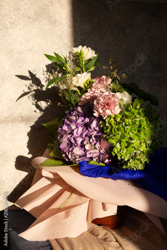 Floral arrangement featuring purple hydrangea, carnation, and freesia flowers in a vase illuminated by direct sunlight, creating dramatic hard light shadows on a neutral stone background.
