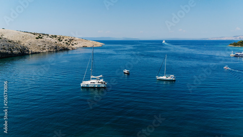 Aerial view of sailboats anchored in the clear blue waters of the Adriatic Sea near the rocky coastline of the island in Rab, Primorje-Gorski Kotar, Croatia.
