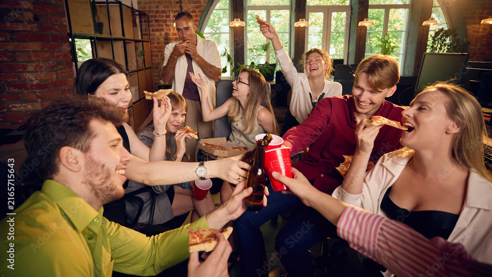 Obraz premium Coworkers toasting with drinks while sharing pizza at office party. Concept of beverage delivery pages, corporate catering services and staff event websites.