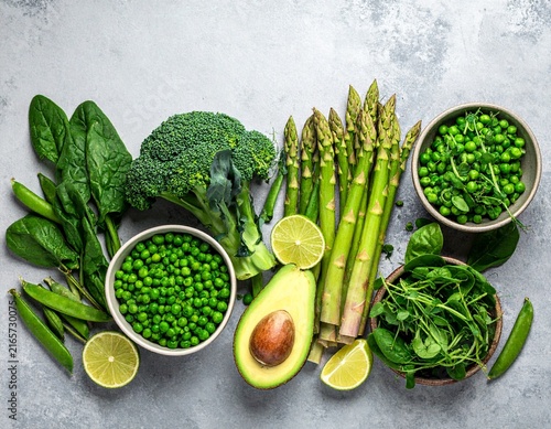 Fresh green vegetables assortment flat lay: broccoli, asparagus, spinach, peas, avocado, lime on grey stone. Healthy vegan food concept, top view.
