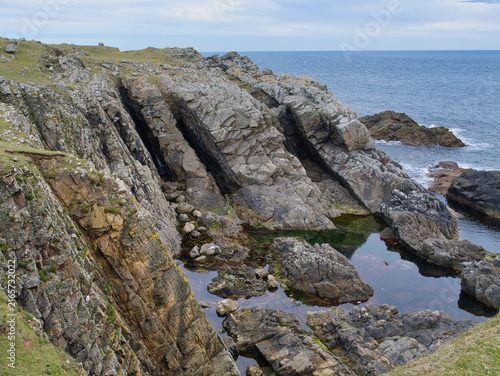 Tilted layers of ancient rock form dramatic coastal cliffs and tide pools at Bu Ness on Fair Isle Shetland UK beneath a pale blue sky