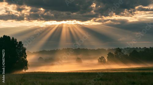 Dramatic sunrise over misty fields, with sunbeams piercing through the clouds.