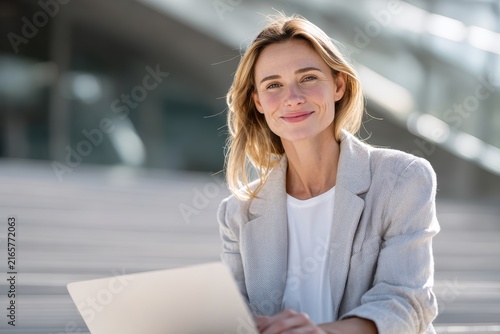 Businesswoman in Light Blazer with Laptop: A confident businesswoman with a warm smile is seated outdoors, holding a laptop on her lap, with modern architecture blurred in the background.