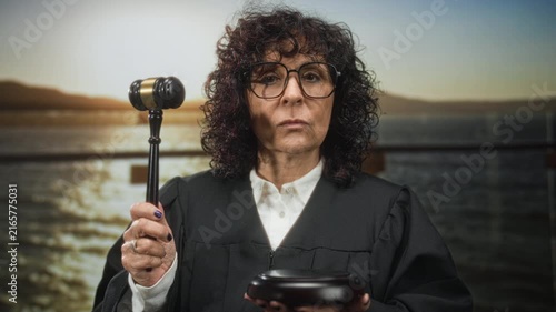 Middleaged caucasian woman judge wearing robe holding gavel poised to strike wooden block in building; solemn authority.