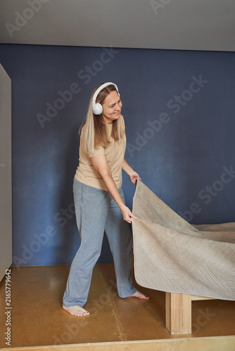 Canvas Print Woman Wearing White Headphones Standing Barefoot Pulling Bedcover Over Mattress In Minimal Bedroom With Blue Wall Wooden Floor And Casual Loungewear