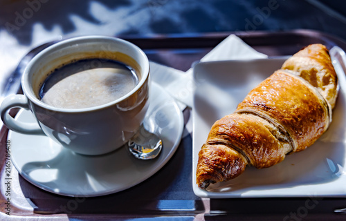 A classic breakfast with a cup of hot coffee with foam in a white bowl and a fresh crispy croissant on a white square plate.