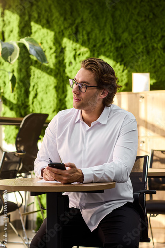 Smiling middle aged man in white shirt sits at round table writing on paper while holding smartphone in bright modern interior with sunlit walls, cabinet and plant