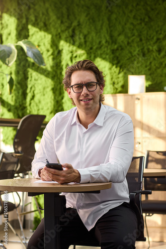 Smiling middle aged man in white shirt sits at round table writing on paper while holding smartphone in bright modern interior with sunlit walls, cabinet and plant