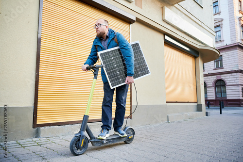 Man rides electric scooter while carrying solar panel. Integration of solar power as sustainable energy source for charging electric scooters, promoting eco-friendly urban transportation.