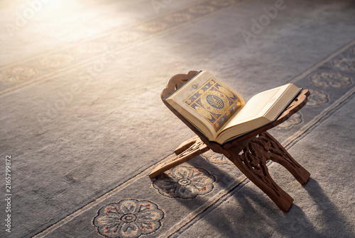 Open holy Quran resting on a wooden stand on a mosque carpet in soft morning sunlight with copy space on a traditional Rihal wooden folding stand