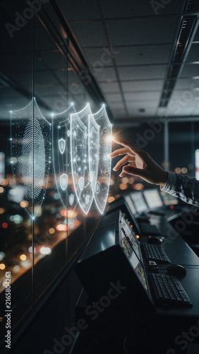 Digital shield icons float in front of a glass window overlooking night city lights as a person reaches toward the glowing holographic interface. Glowing lines form protective patterns over an urban