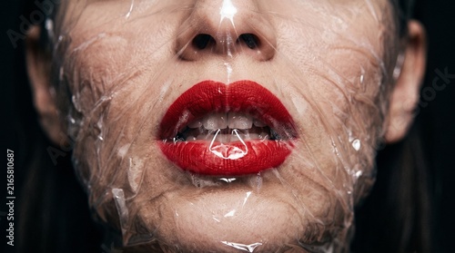 Close-up of a person's face with bright red lips covered tightly by clear plastic wrap, creating a distorted and unsettling effect.