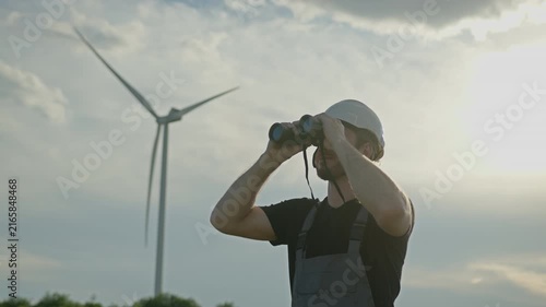Man in hard hat and overalls uses binoculars to observe wind turbine against cloudy sky, representing renewable energy sector. Engineer monitoring sustainable technology infrastructure