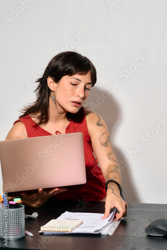 A focused woman works at her desk with laptop and papers. She’s deeply concentrated. Picking up business papers while holding her laptop at office