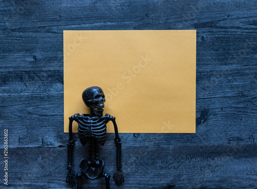 Halloween concept photo of yellow blank paper with a black plastic skeleton flat lay background.