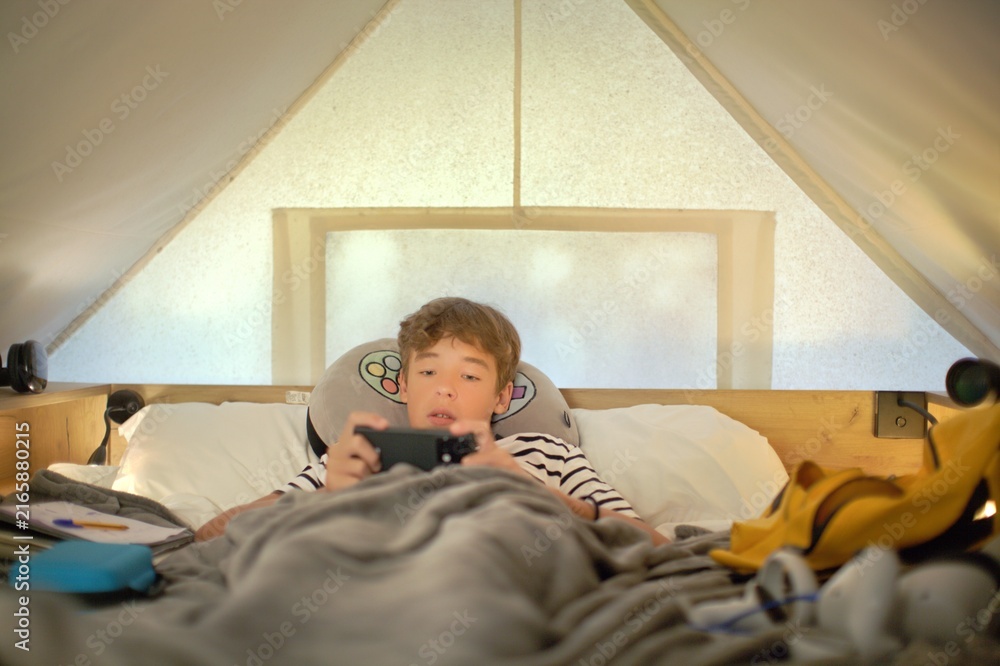 Fototapeta premium Teenage boy lies under a blanket in a glamping tent and holds a phone with both hands, focused on a mobile game, soft daylight through the canvas walls