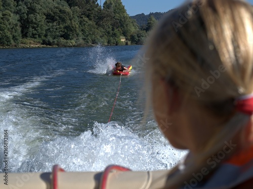 Картината върху платно Boy rides a red towable tube through the wake of a speeding boat, blonde woman w