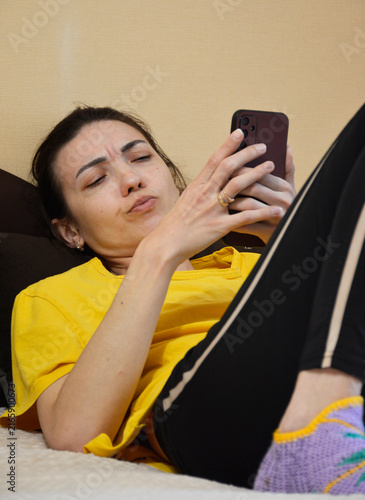 A young, beautiful woman is lying on the sofa with a phone in her hands. She is looking at the smartphone screen and frowning with dissatisfaction. negative emotions are visible on her beautiful face